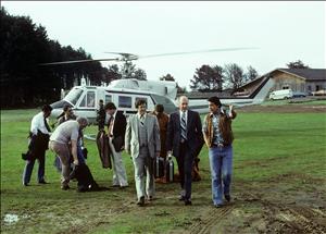 Two men in suits and ties walk away from a helicopter beside a man in a vest and jeans who gestures ahead of them. Other people stand around behind them. The helicopter has landed on a patch of grass with wooden buildings and a forested hillside in the distance. 